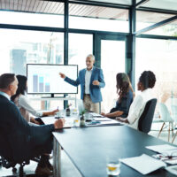 A business meeting in a bright conference room, where a man in a blue blazer presents a bar graph on a screen to engaged colleagues seated around a table.
